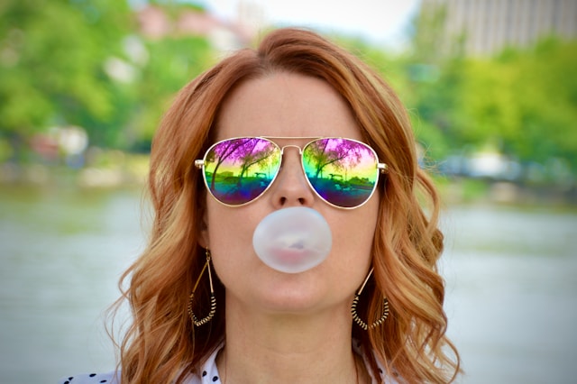 Image of a woman in sunglasses blowing a bubble with bubble gum.